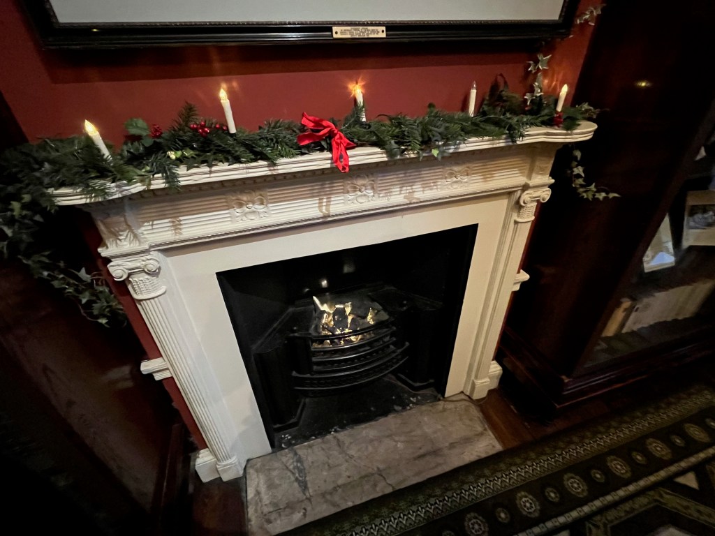 A fireplace in the Charles Dickens Museum with a white frame, the mantlepiece of which is lined with decorative green foliage supporting five candles, and a red bow tied around the centre of it.