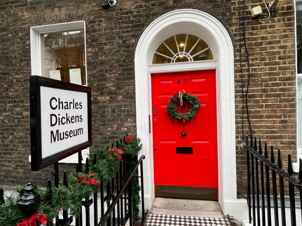 A white sign with large black text saying Charles Dickens Museum stands next to a short path leading to the vivid red front door, on which a green wreath with a decorative gold bow hangs just below the black number 49 at the top.