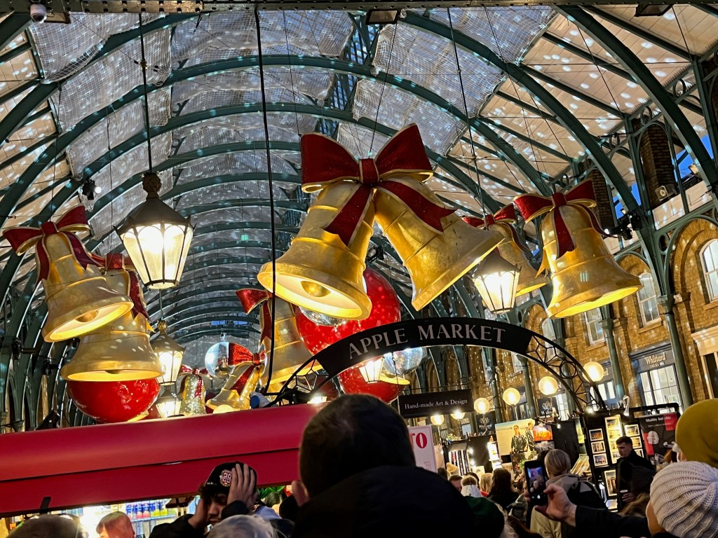 Christmas decorations hanging from the arched ceiling inside Covent Garden market, including large gold bells with glittering red bows on the top, large red baubles and mirror balls.