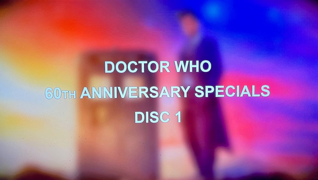 Audio navigation screen from the Doctor Who 60th Anniversary Specials Blu-ray. Over a blurred image of the Doctor standing next to the Tardis against a colourful background, large white uppercase text reads Doctor Who 60th Anniversary Specials Disc 1.