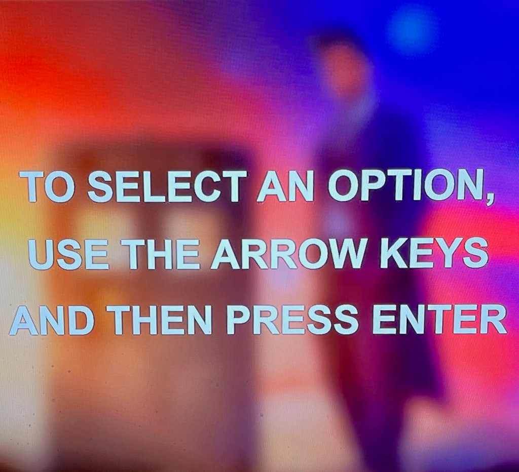 Audio navigation screen from the Doctor Who 60th Anniversary Specials Blu-ray. Over a blurred image of the Doctor standing next to the Tardis against a colourful background, large white uppercase text reads To select an option, use the arrow keys and then press Enter.