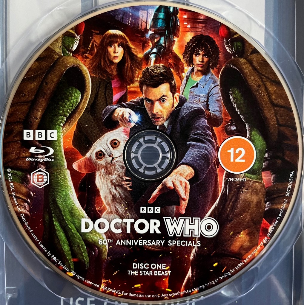 Disc 1 in the Blu-ray steelbook for the Doctor Who 60th Anniversary Specials, containing The Star Beast. The colourful artwork shows various characters from the episode. Front and centre is David Tennant's Doctor, who is reaching towards us with his left hand while pointing his sonic screwdriver with the other. Around him are Donna Noble, her daughter Rose, the Meep and a Wrarth Warrior.