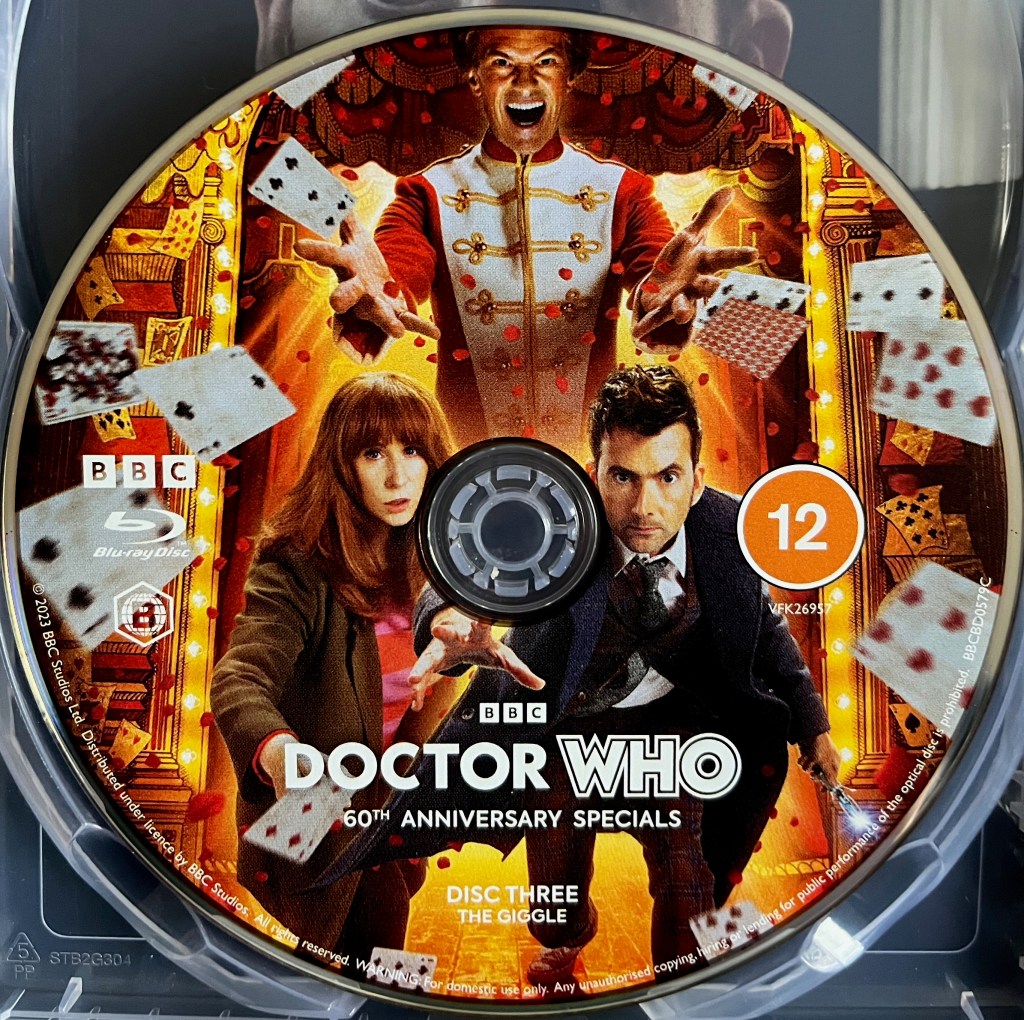 Disc 3 in the Blu-ray steelbook for the Doctor Who 60th Anniversary Specials, containing The Giggle. The colourful artwork shows David Tennant's Doctor and Donna Noble running urgently towards us, while the Toymaker towers over them in the background, standing on a brightly lit stage and laughing as he throws playing cards everywhere.