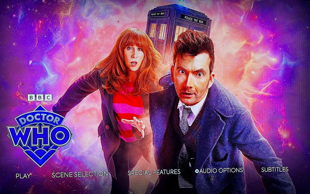 The main menu on the Blu-ray for the Doctor Who 60th Anniversary Specials, showing David Tennant's Doctor and Donna Noble running urgently towards us, while the Tardis flies through the universe in the background. The Doctor Who logo is in the bottom left corner.