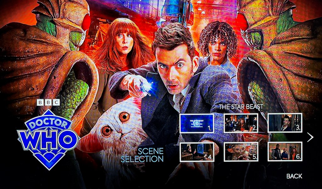 Scene Selection menu on the Blu-ray disc for The Star Beast, in the box set for the Doctor Who 60th Anniversary Specials. The colourful background shows various characters from the episode. Front and centre is David Tennant's Doctor, who is reaching towards us with his left hand while pointing his sonic screwdriver with the other. Around him are Donna Noble, her daughter Rose, the Meep and a Wrarth Warrior. Overlaid in the bottom right of the screen are 6 clickable images representing different points in the episode. The Doctor Who logo is in the bottom left corner.