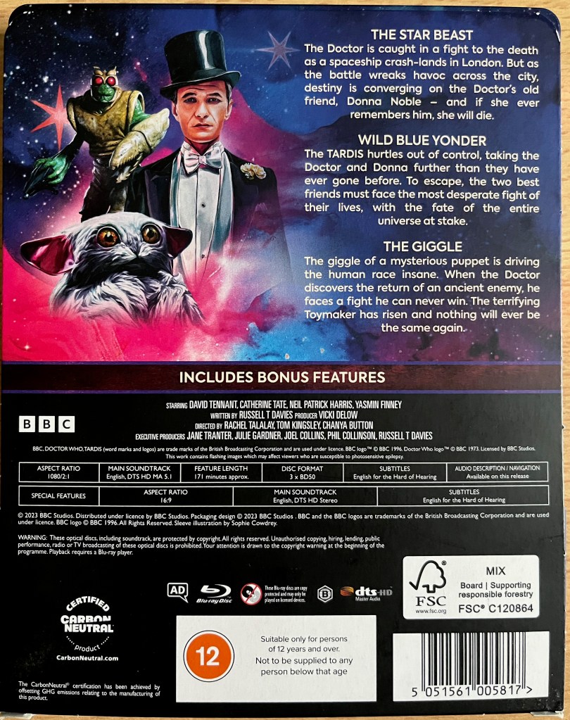 The back cover of the cardboard pocket that houses the steelbook for the Doctor Who 60th Anniversary Specials. It shows an image of a green alien Wrath Warrior, the small white furry Meep and the smartly suited Toymaker alongside descriptions of the 3 episodes. Below that is a line saying that the set includes bonus features, without specifying what they are, followed by credits and technical specifications.