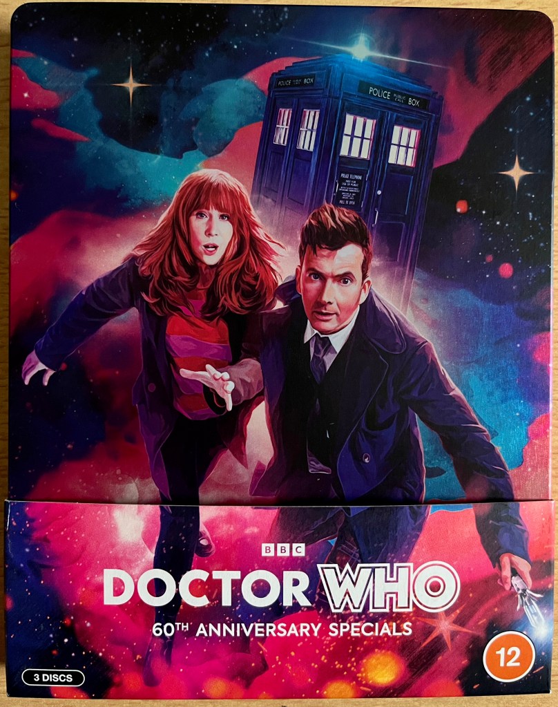 The front cover of the steelbook for the Doctor Who 60th Anniversary Specials, which is sitting in a cardboard pocket that has the Doctor Who logo on it. The cover shows David Tennant's Doctor and Donna Noble running urgently towards us, while the Tardis flies through the universe in the background.