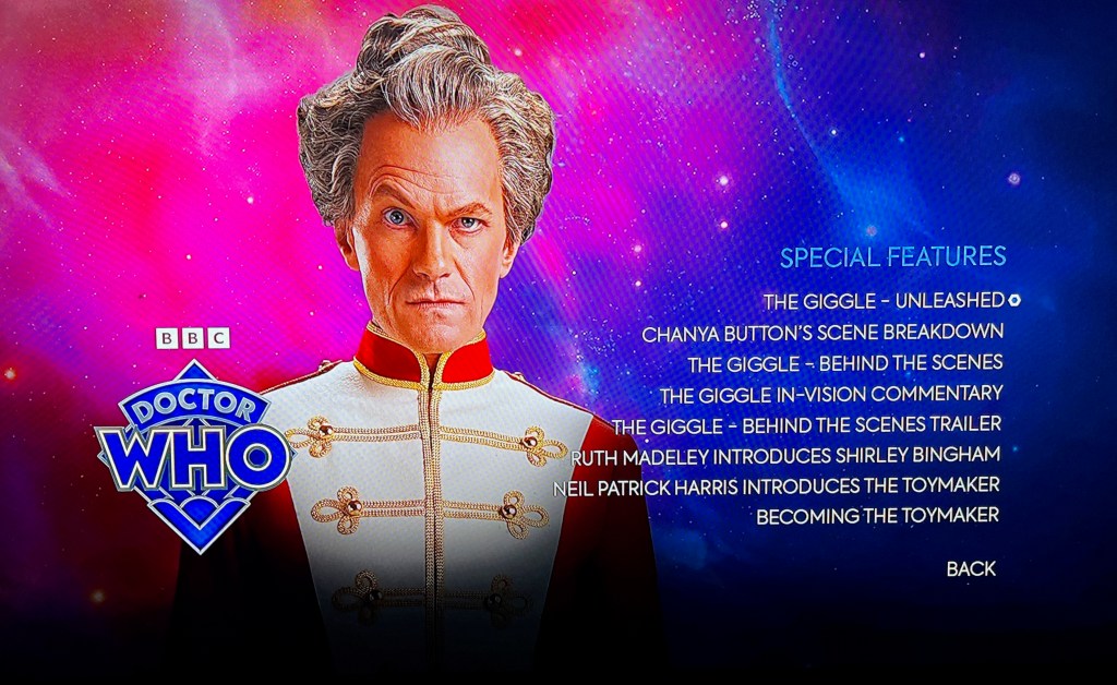 Special Features menu for The Giggle, in the Doctor Who 60th anniversary Specials Blu-ray. The menu shows the options to choose from, alongside the Toymaker in a white top with red sleeves and collar and gold detailing. The Doctor Who logo is in the bottom left corner.