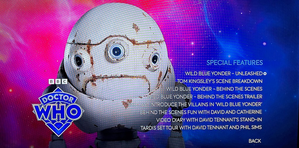 Special Features menu for Wild Blue Yonder, in the Doctor Who 60th anniversary Specials Blu-ray. The menu shows the options to choose from, next to the large round head of a white robot that is covered in patches of brown rust. The Doctor Who logo is in the bottom left corner.