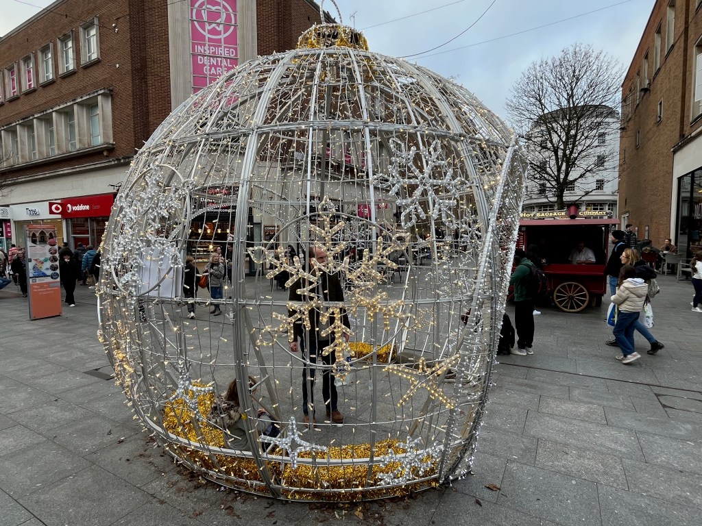Side view of the large bauble-shaped structure on the High Street in Exeter that people can walk through, decorated with gold and silver snowflakes, and covered in lots of small lights.