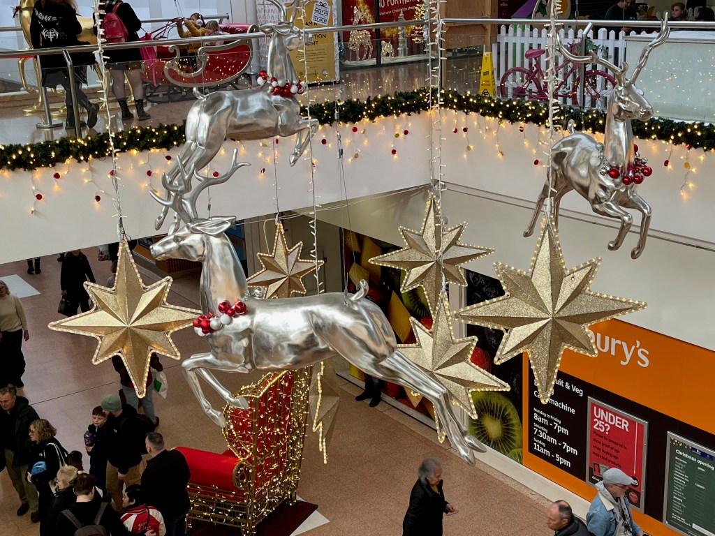 Silver reindeer and large gold stars with lights around the edges hanging from the ceiling in Exeter's Guildhall Shopping Centre. On the ground floor below, a red throne with festive lights all over the back and sides is available for people to sit in for photos.