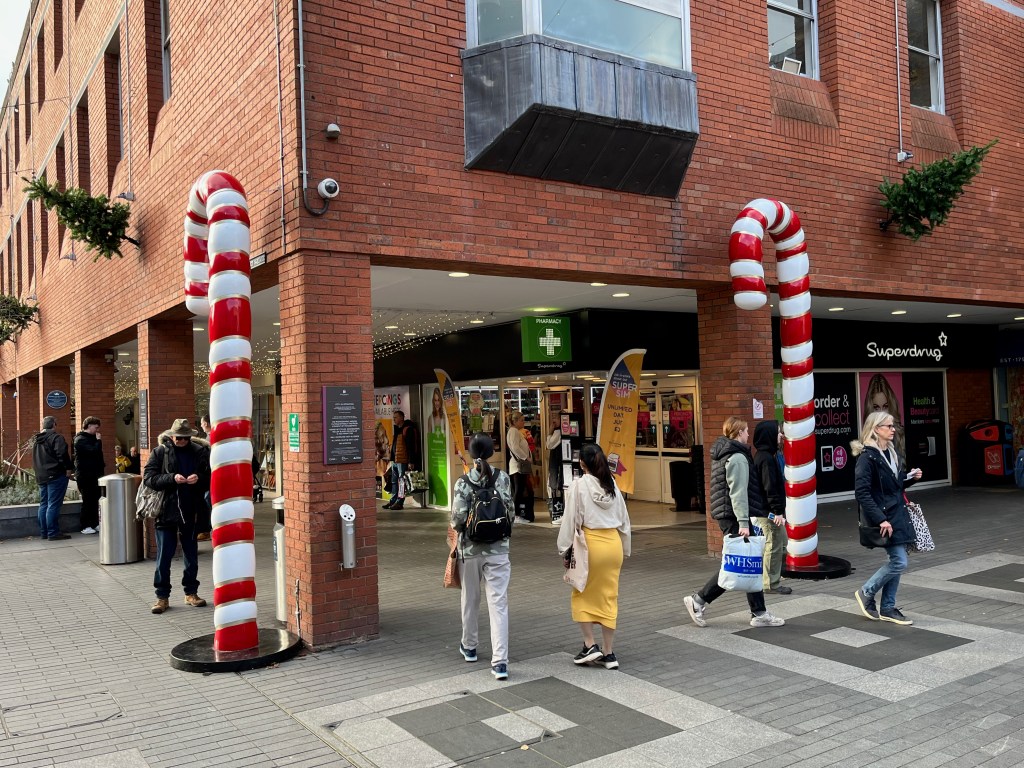 Two very tall red and white striped candy canes standing in an outdoor area of Exeter's Guildhall Shopping Centre, by the entrance to a corridor that leads to the inside of the shopping centre.
