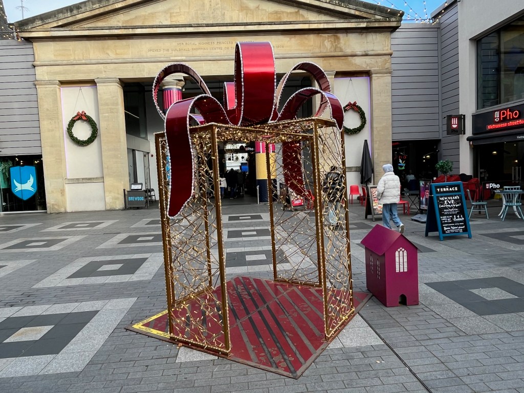 A tall box with open corners that people can walk through, featuring a vertical wall of lights in the centre on each side, and a large red bow with lit edges on the roof.