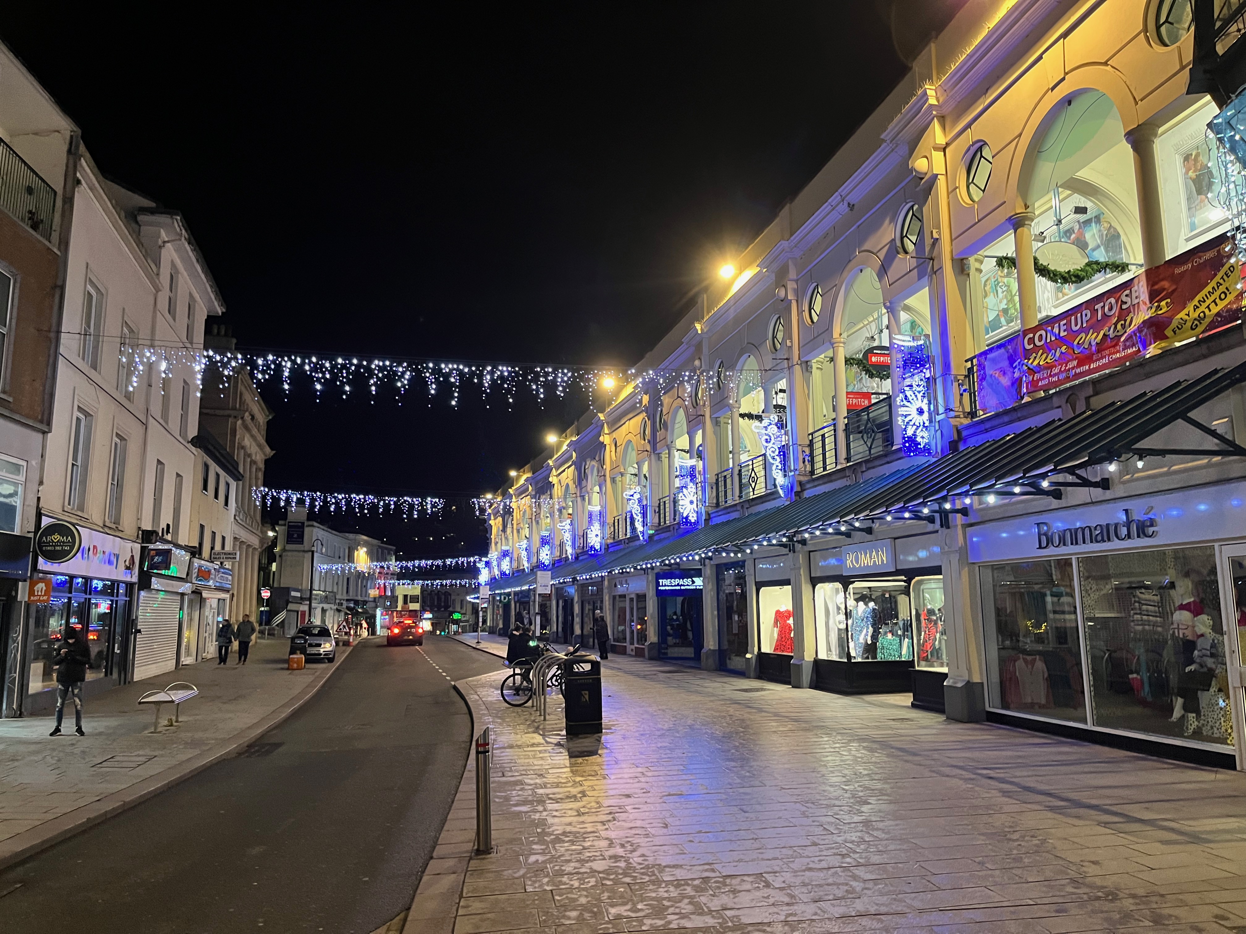 Strings of festive lights hanging over Fleet Street in Torquay, along with some lit snowflakes on the first floors of the buildings on the right hand side, above the shops on the ground floor.
