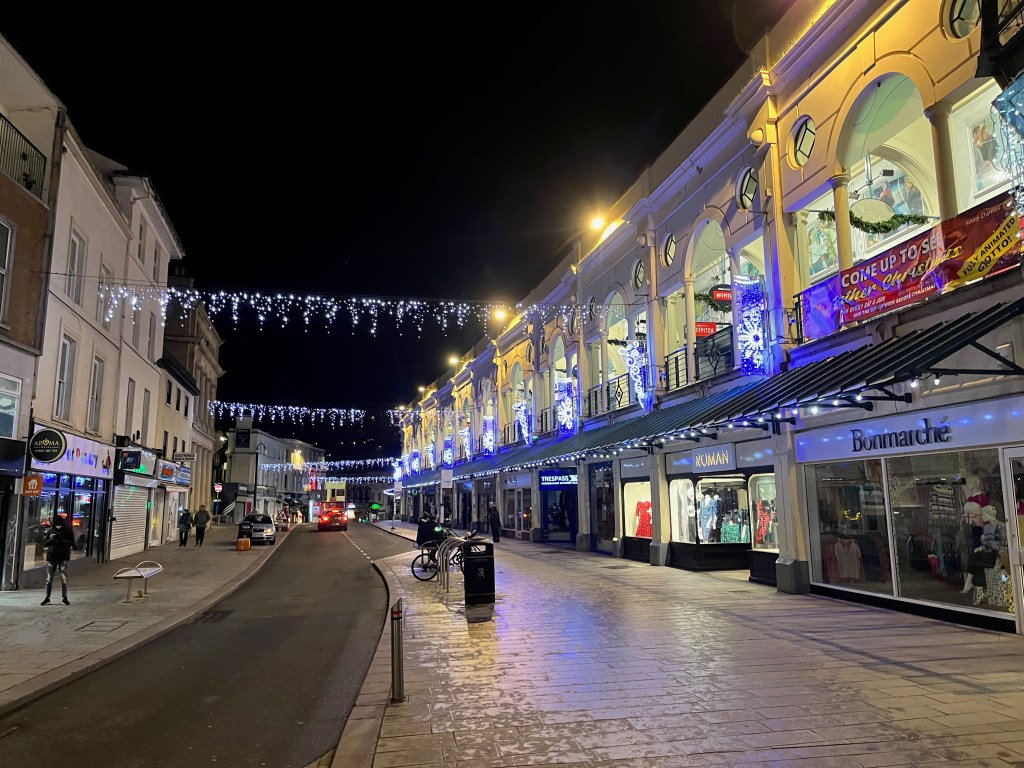 Strings of festive lights hanging over Fleet Street in Torquay, along with some lit snowflakes on the first floors of the buildings on the right hand side, above the shops on the ground floor.