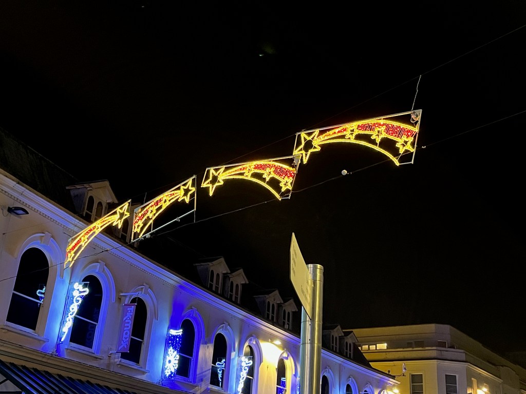 Christmas lights over Fleet Street in Torquay, designed to look like comets with a bright yellow outline. There are 3 small stars along the tail section of each comet, and a big star at the front.