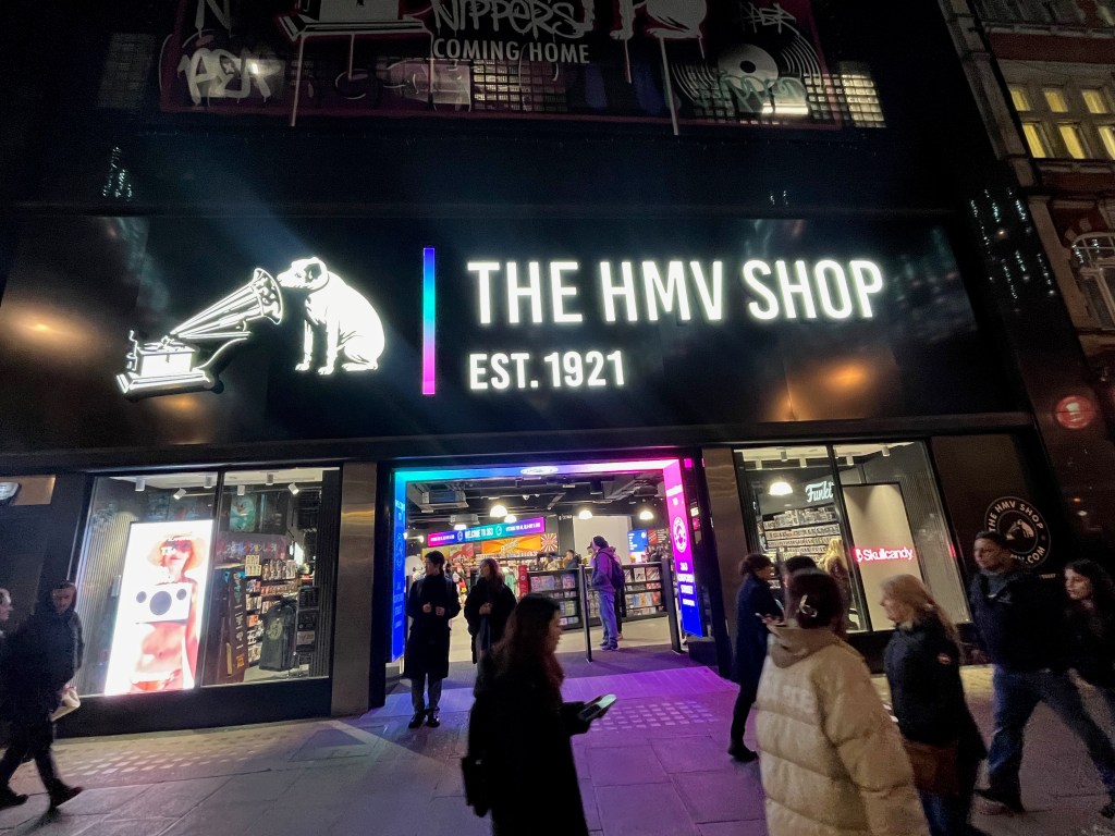 The HMV shop in Oxford Street. The large sign above the doorway glows white on its black background, showing the iconic image of a dog looking into a gramophone, next to the words The HMV Shop, established 1921.
