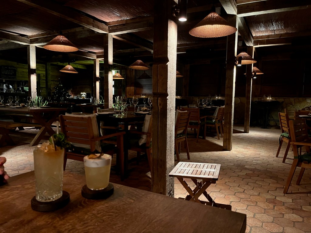 The In Horto restaurant, with tables set amongst set in between tall pillars under atmospheric lighting. Two cocktails are on our table.