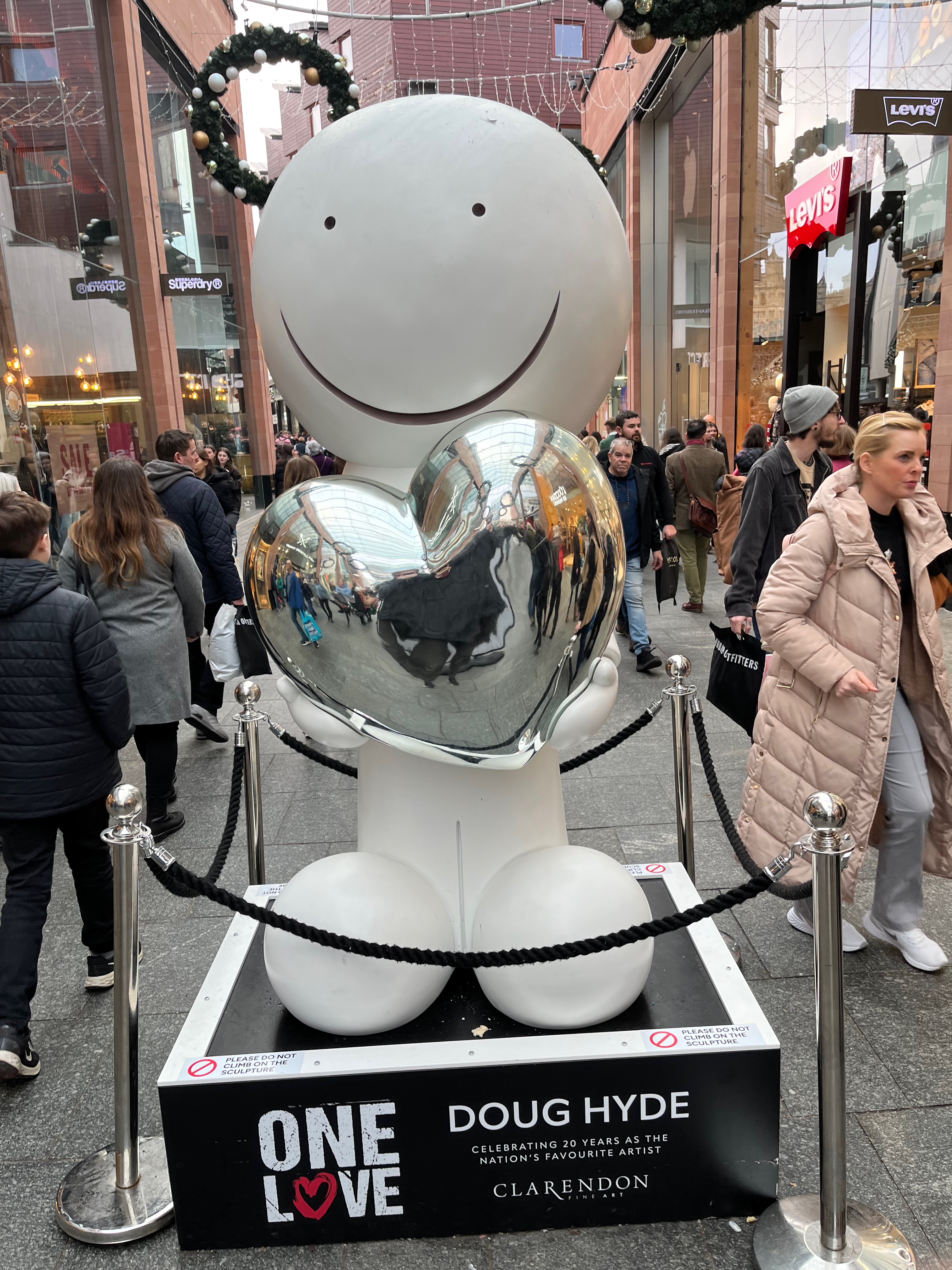 A large sculpture of a white figure with bulbous feet and a big round head, which simply has a black line drawn in a big smile and 2 black dots for eyes. The figure is holding a large silver heart, that is reflecting the people walking around him.