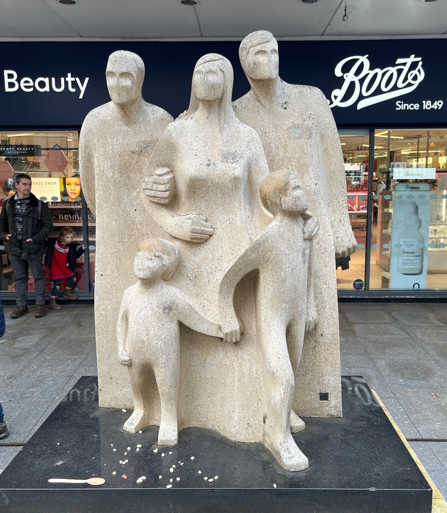 The Pedestrian sculpture outside Boots on Exeter High Street, showing 3 adults and 2 children. The figures are all linked together, as if slightly morphing into each other, and their faces have minimal detailing, with simple hairstyles and indentations for eye sockets.