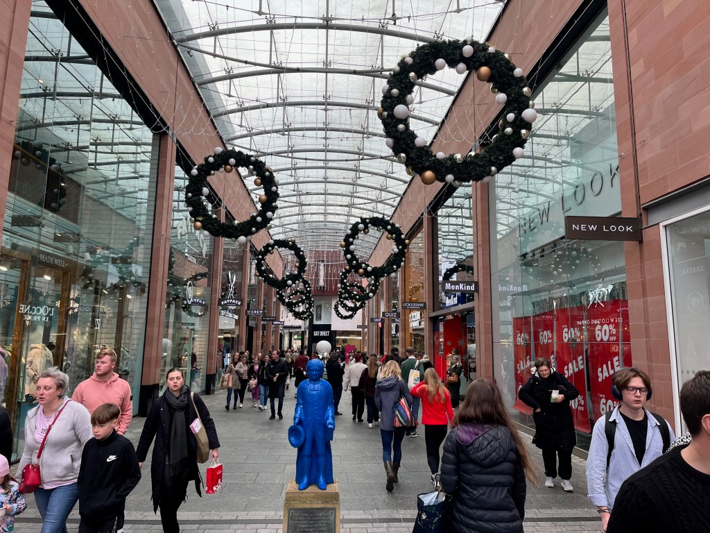 Princesshay shopping centre in Exeter. Large wreaths, covered in white, silver and gold baubles, hang at regular intervals from the arched glass ceiling over the corridor, above the shoppers and the Blue Boy statue.