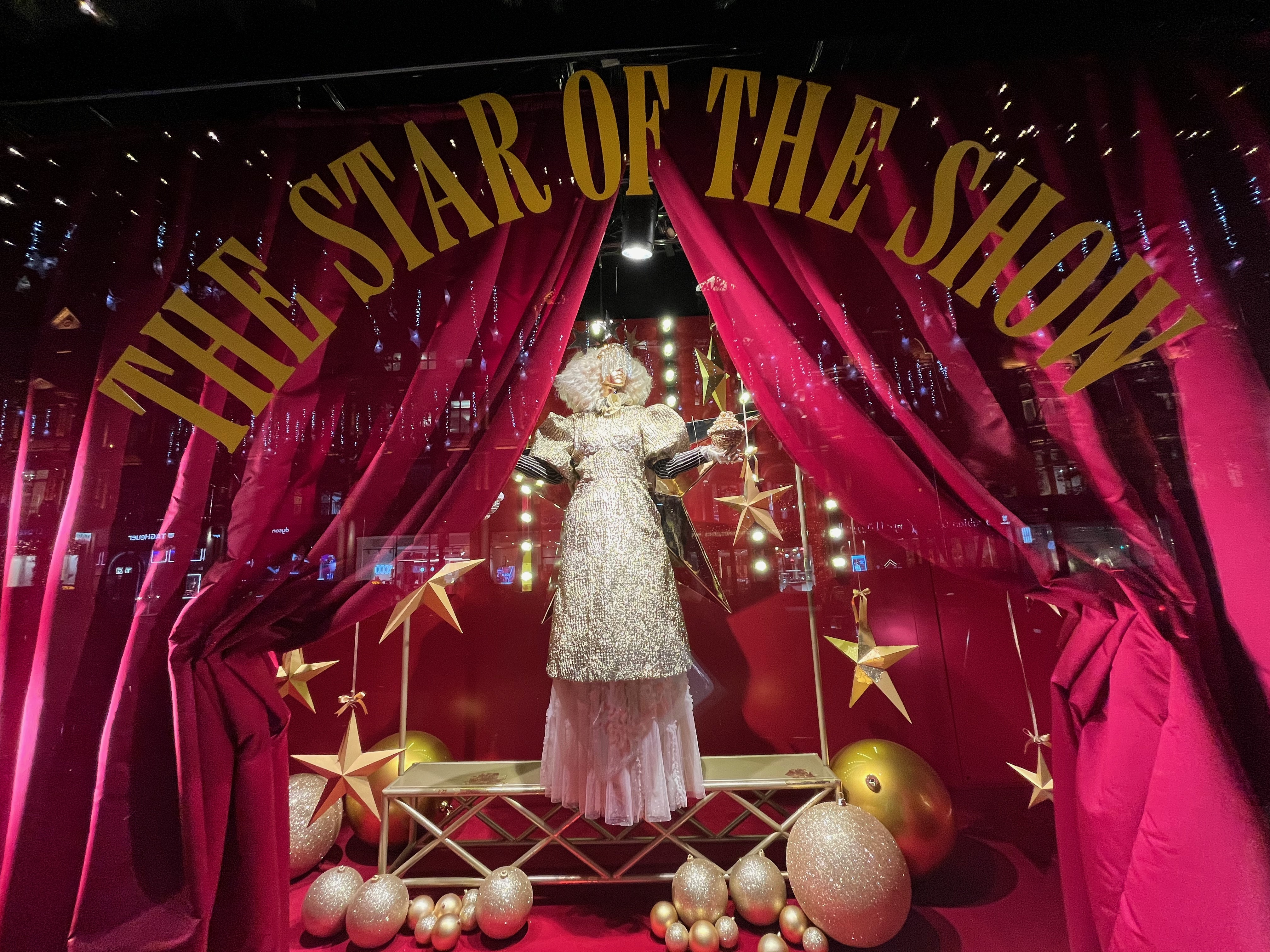 Selfridges window display, showing a mannequin wearing a long glittering dress and a white wig. On the window itself is large yellow text that says The star of the show, and it forms an arch through which you can see the view.