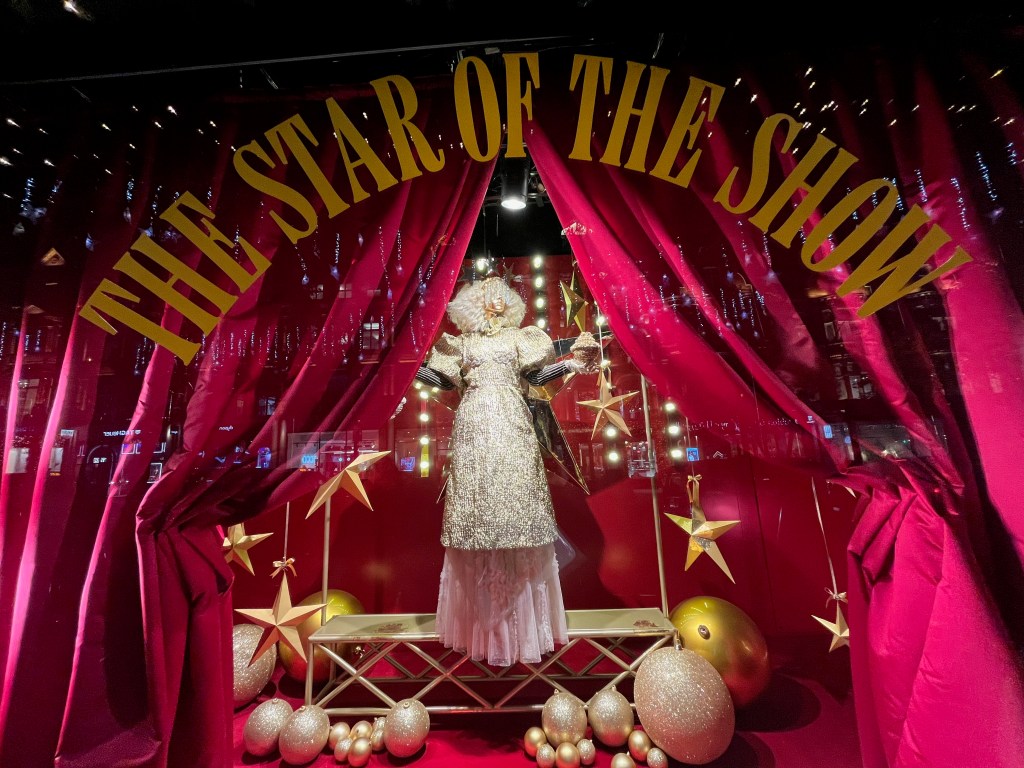 Selfridges window display, showing a mannequin wearing a long glittering dress and a white wig. On the window itself is large yellow text that says The star of the show, and it forms an arch through which you can see the view.
