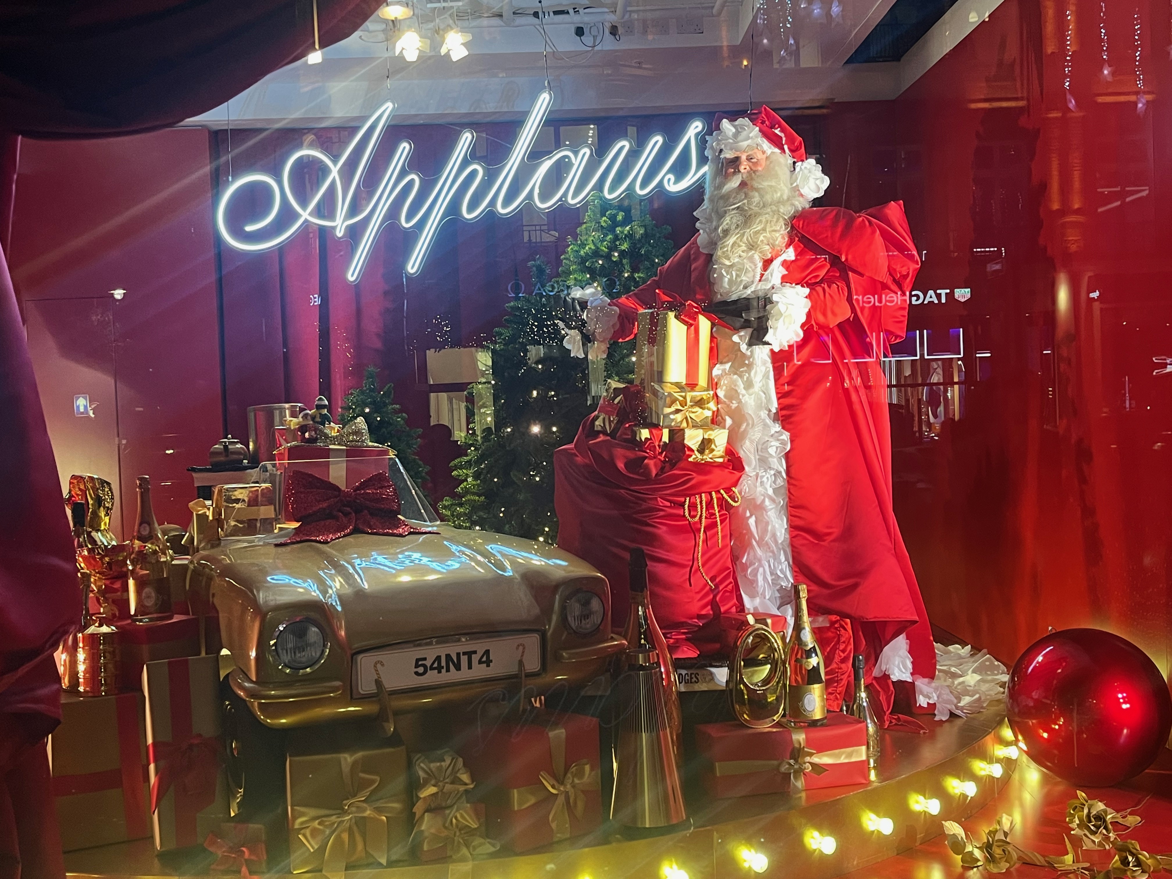 Selfridges window display, showing Santa Claus unloading a large sack of presents, while standing beside a car also filled with gifts, which has the registration number 54NT4. A bright sign behind him has the word Applause lit up in handwriting style lettering.