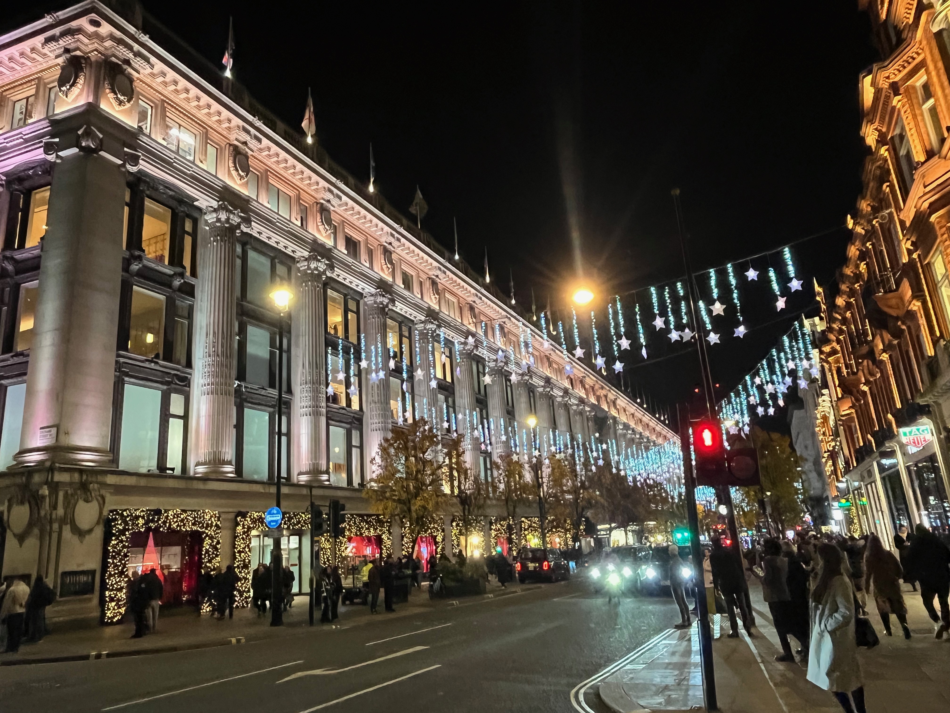 View across Oxford Street to Selfridges. Cables are suspended high across the street, hanging from which are bright stars on lit-up wires of varying lengths.