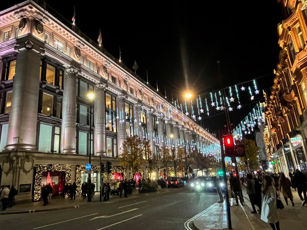 View across Oxford Street to Selfridges. Cables are suspended high across the street, hanging from which are bright stars on lit-up wires of varying lengths.