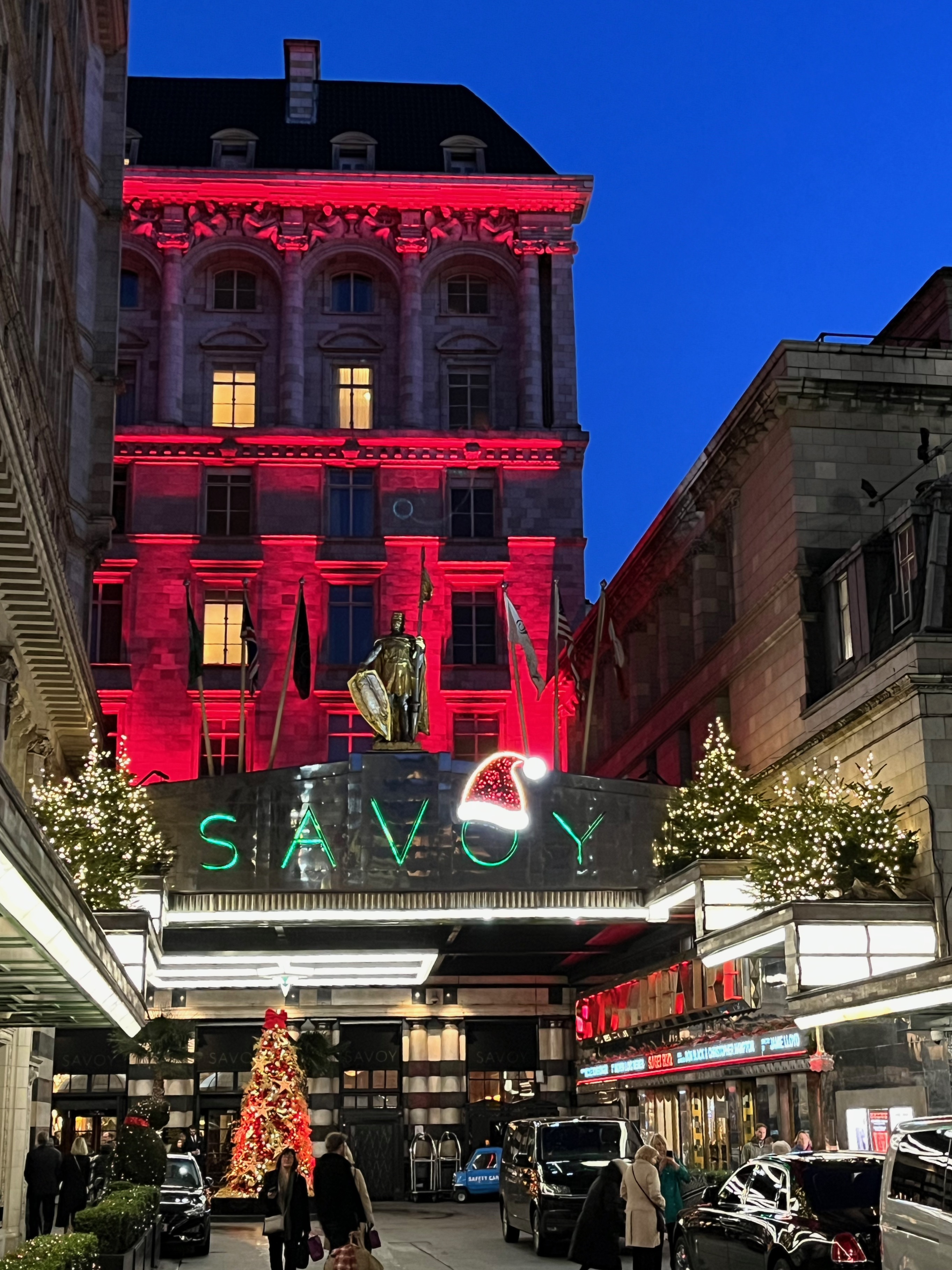 Christmas lights over the entrance to the Savoy hotel. There is a lit Santa hat on the O of the word Savoy, while there are brightly lit Christmas trees each side of that sign. The tall building behind the sign is lit up in red.