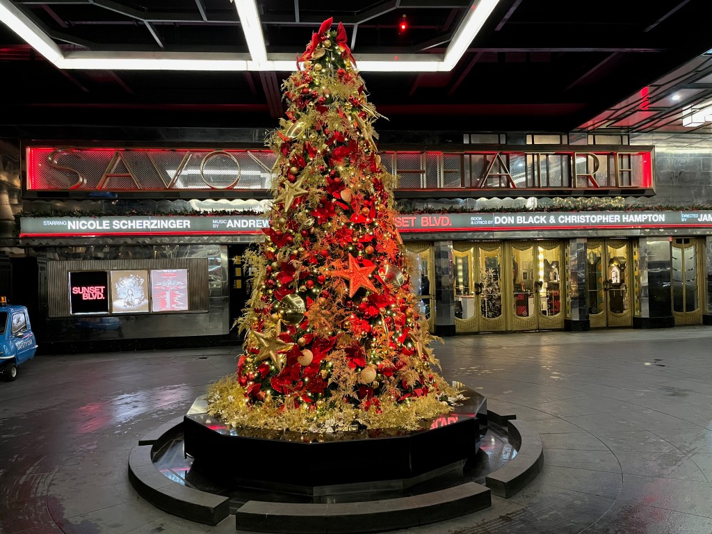 Large Christmas tree on a round base outside the entrance to the Savoy hotel, mainly in red, green and gold colours, adorned with glittering stars and baubles as well as lights.