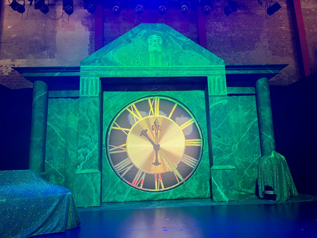 The stage for The Time Machine at the Park Theatre. A large image of a clock, with gold Roman numerals on a black circle around a gold centre, is on a wall between two columns that are holding up a triangular canopy. The set is bathed in green light. 