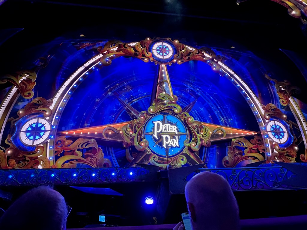 The stage for the Peter Pan panto, revealing a large 4-point compass style design against a blue backdrop. Only the North, West and East points are visible, the South point hidden below stage level. The arms of the compass star that span the entire stage each have a few lights along their length. In the centre is a smaller circular compass, surrounded by a frame of ornate swirling shapes, with the words Peter Pan in big white letters in the middle.