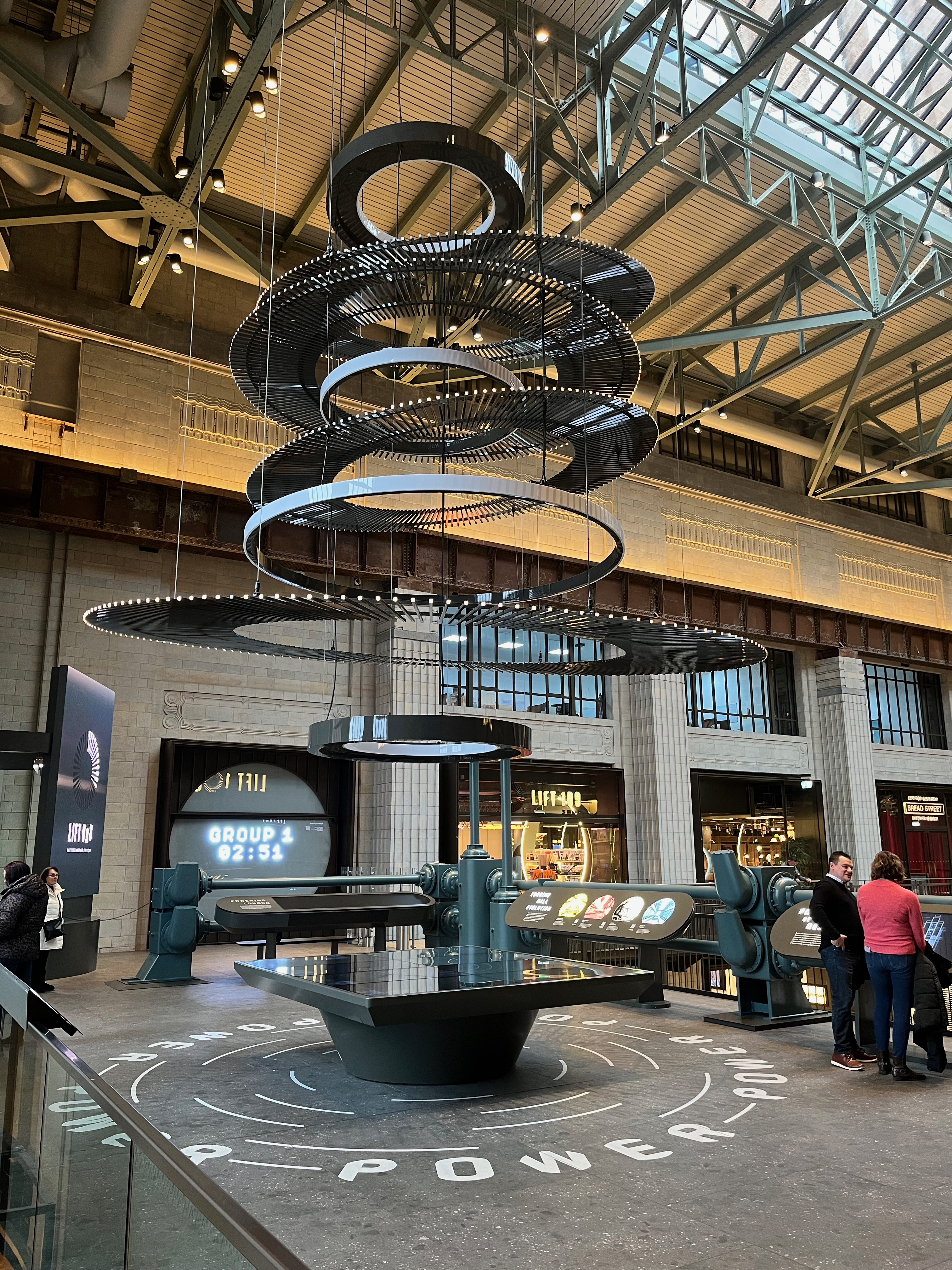 A tall metal spiral-shaped sculpture, hanging above the centre of a small exhibition area for Lift 109 in Battersea Power Station, which has a black interactive screen table in the centre, and other interactive panels around the edge of the space. On a wall at the back, a countdown is in progress, so that visitors know when they will be moving into the next area.
