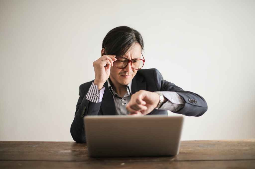 A businessman looking at his watch as he sits in front of a laptop.