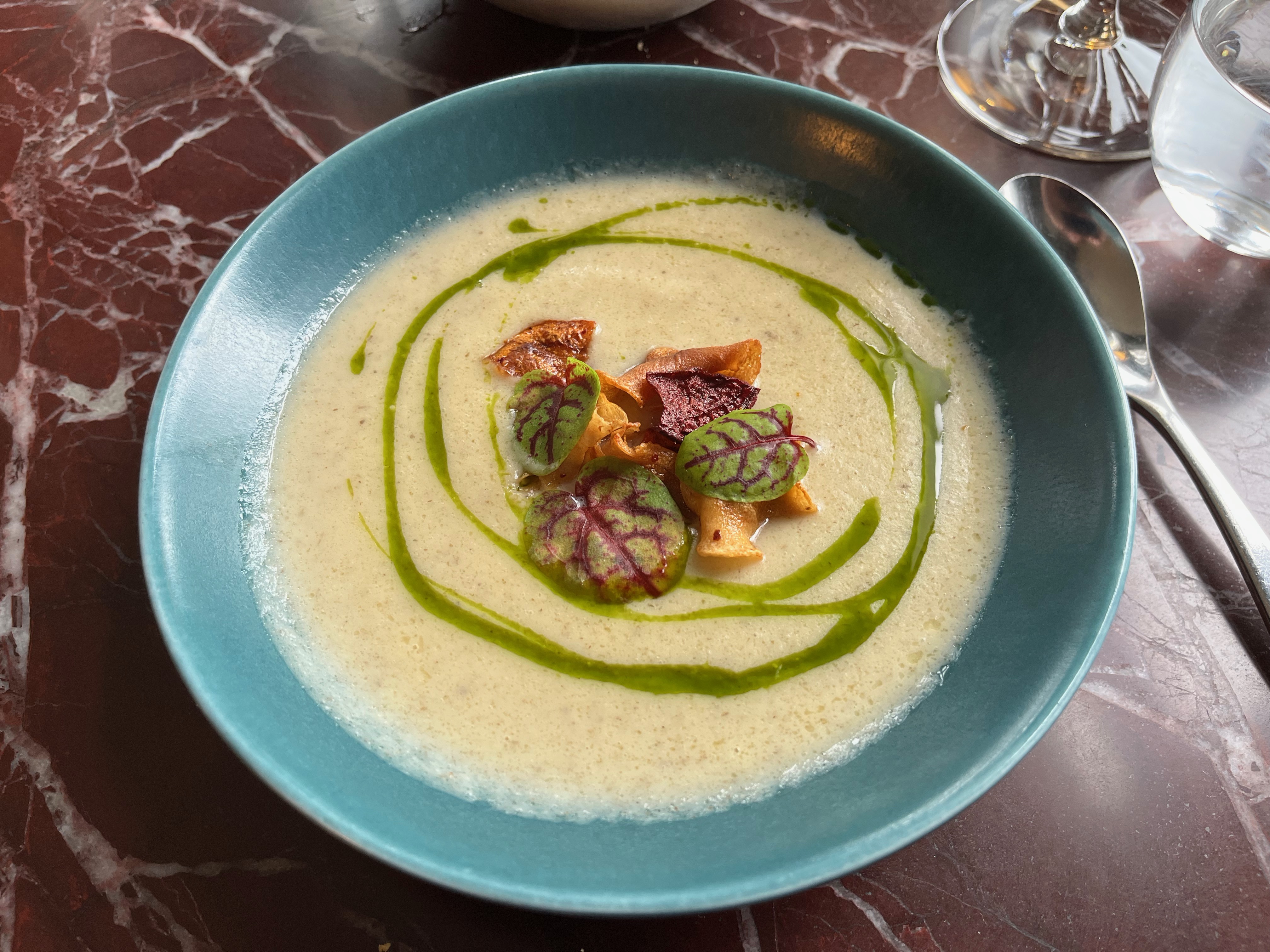 A bowl of artichoke soup, a very light green colour with a circular swirl of much darker green, and leafy decorations in the centre.