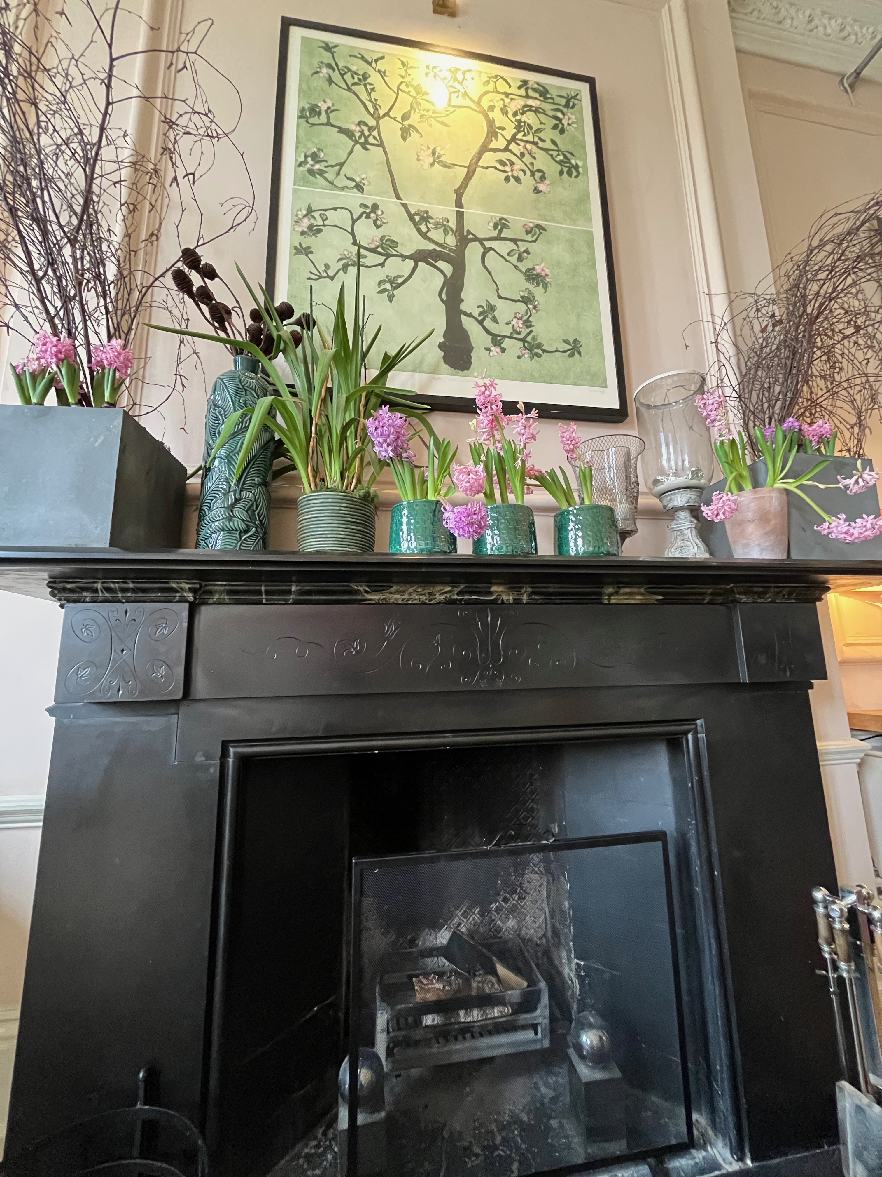 Decor on the walls in the Bingham Riverhouse. An artistic portrait of a tree with flowers on some of its many branches hangs over a fireplace mantlepiece, on which are several small pots with pink and purple flowers and greenery.