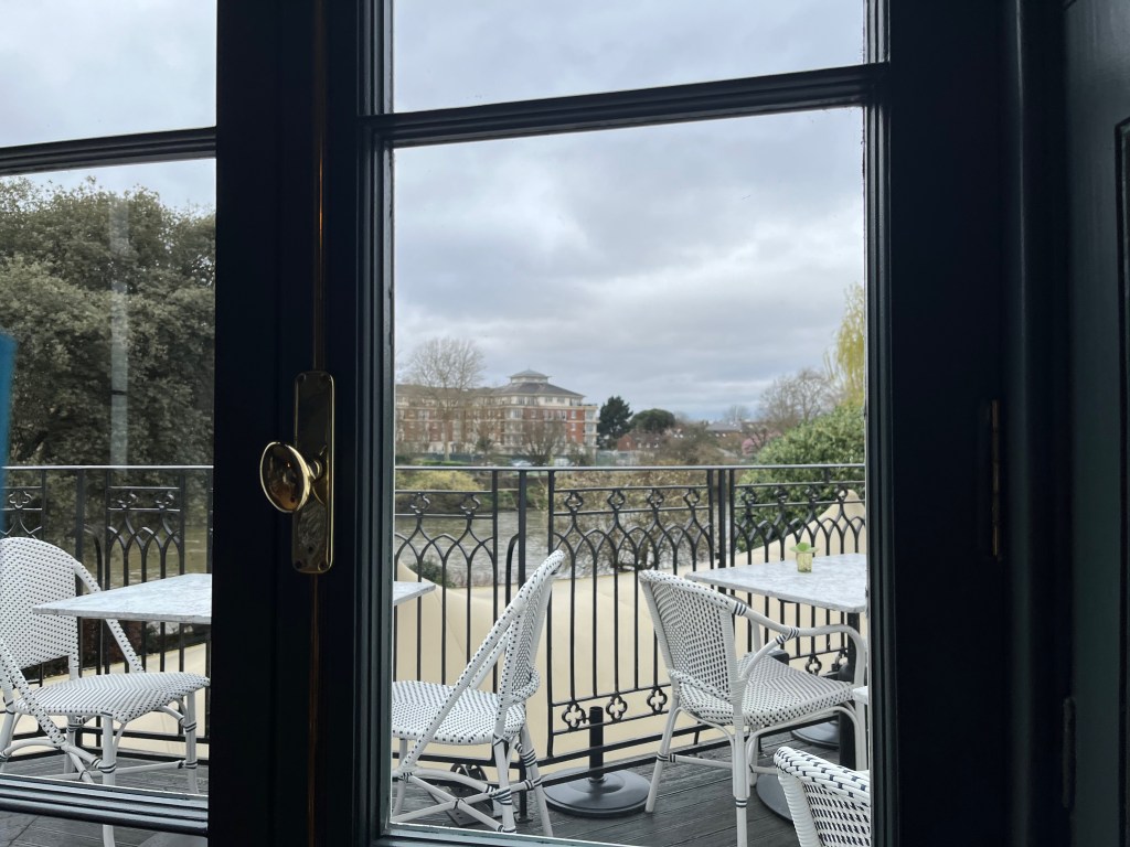 View through the window of the Bingham Riverhouse restaurant, showing tables and chairs in an outdoor seating area that overlooks the River Thames.