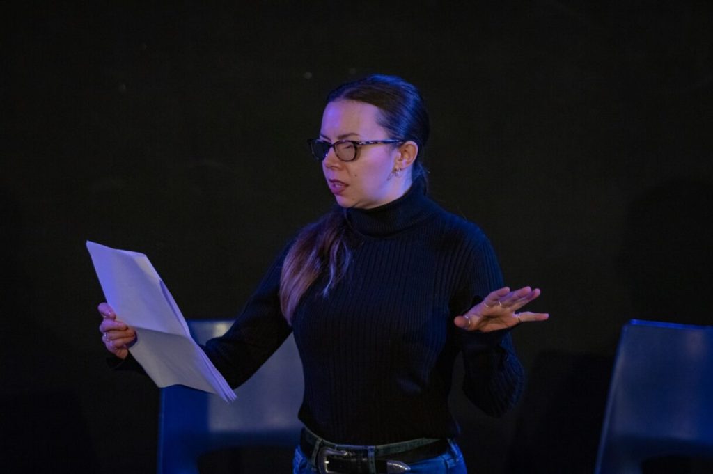 Georgie Morrell, a white woman with long brown hair, wears glasses and a black jumper, and holds a script.