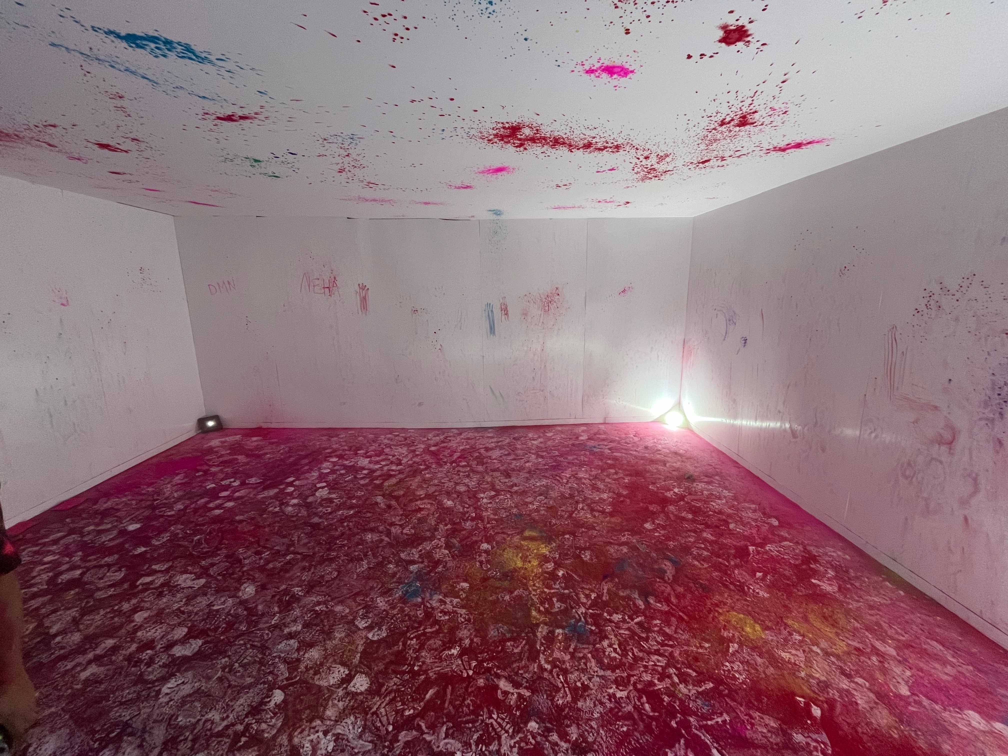 The empty House of Holi room, showing the aftermath of the paint throwing session. The floor is covered in colour, mainly red but with pink, yellow and green mixed in, and a lot of white footprints too. There are lots of splashes of colour on the walls and ceiling as well.