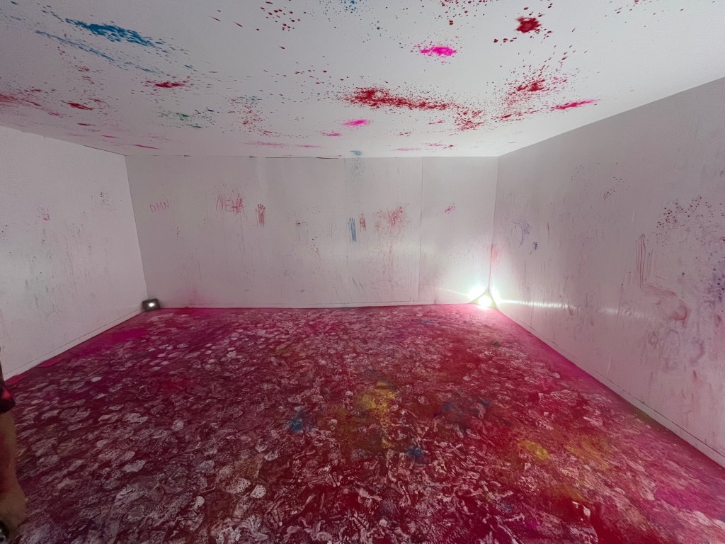 The empty House of Holi room, showing the aftermath of the paint throwing session. The floor is covered in colour, mainly red but with pink, yellow and green mixed in, and a lot of white footprints too. There are lots of splashes of colour on the walls and ceiling as well.