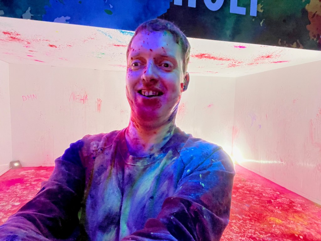 Glen smiling while covered in various colours of powder paint, in front of the messy House of Holi room.