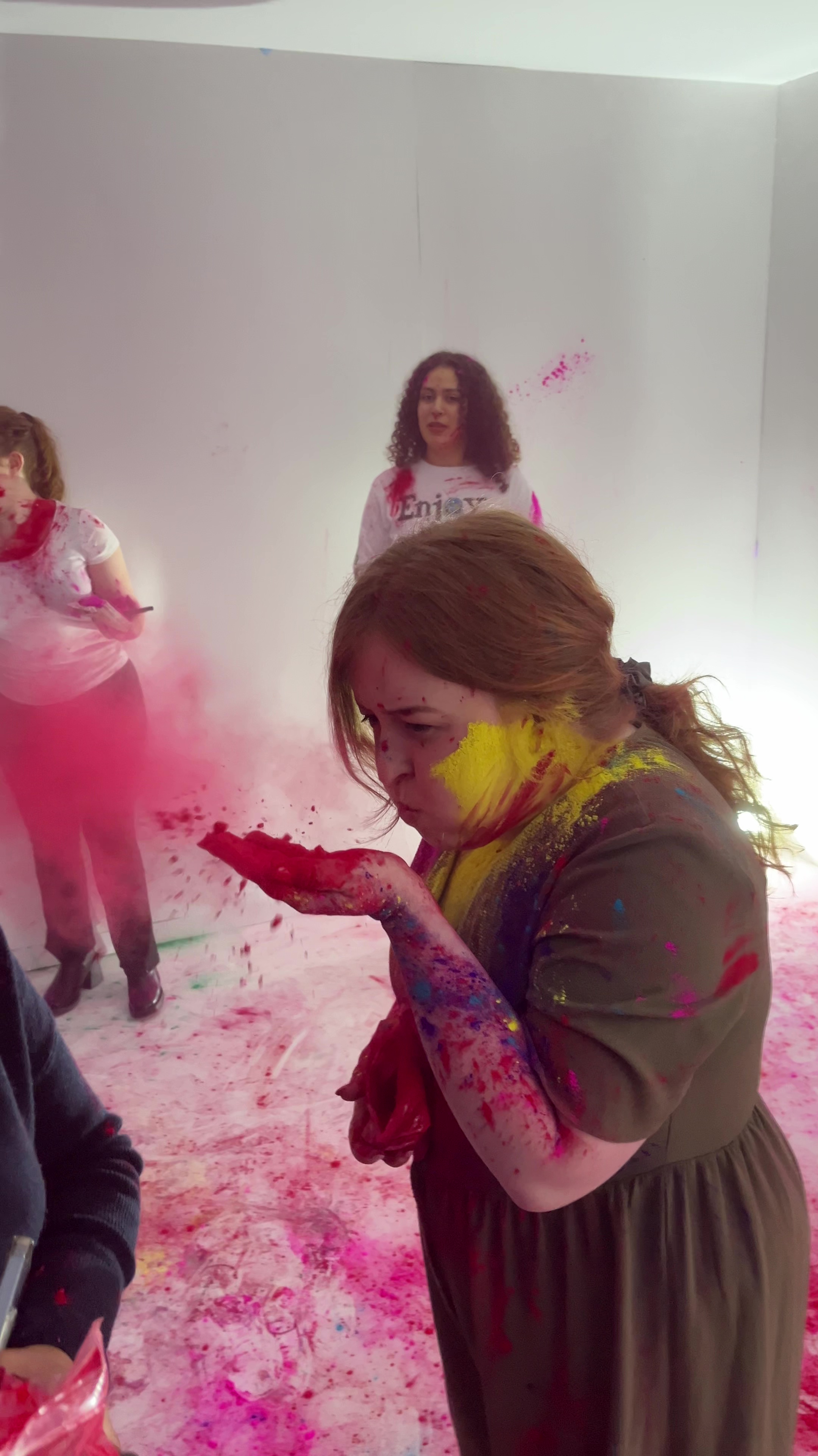Emily Davison in the House of Holi room. She is covered in different colours of powder paint, and is blowing red powder from her hand to make a cloud in front of her.
