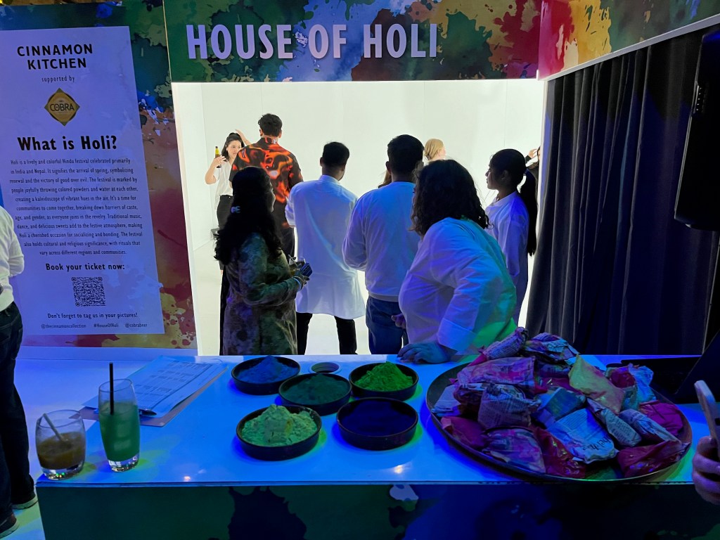 People gathered at the entrance of a large white cube-shaped room. The outer wall around the entrance is covered in colour, as if paint has been splashed all over it. White capital letters against this background, over the top of the entrance, says House Of Holi. A tall sign to the wall on the left of the entrance explains what the festival is about.