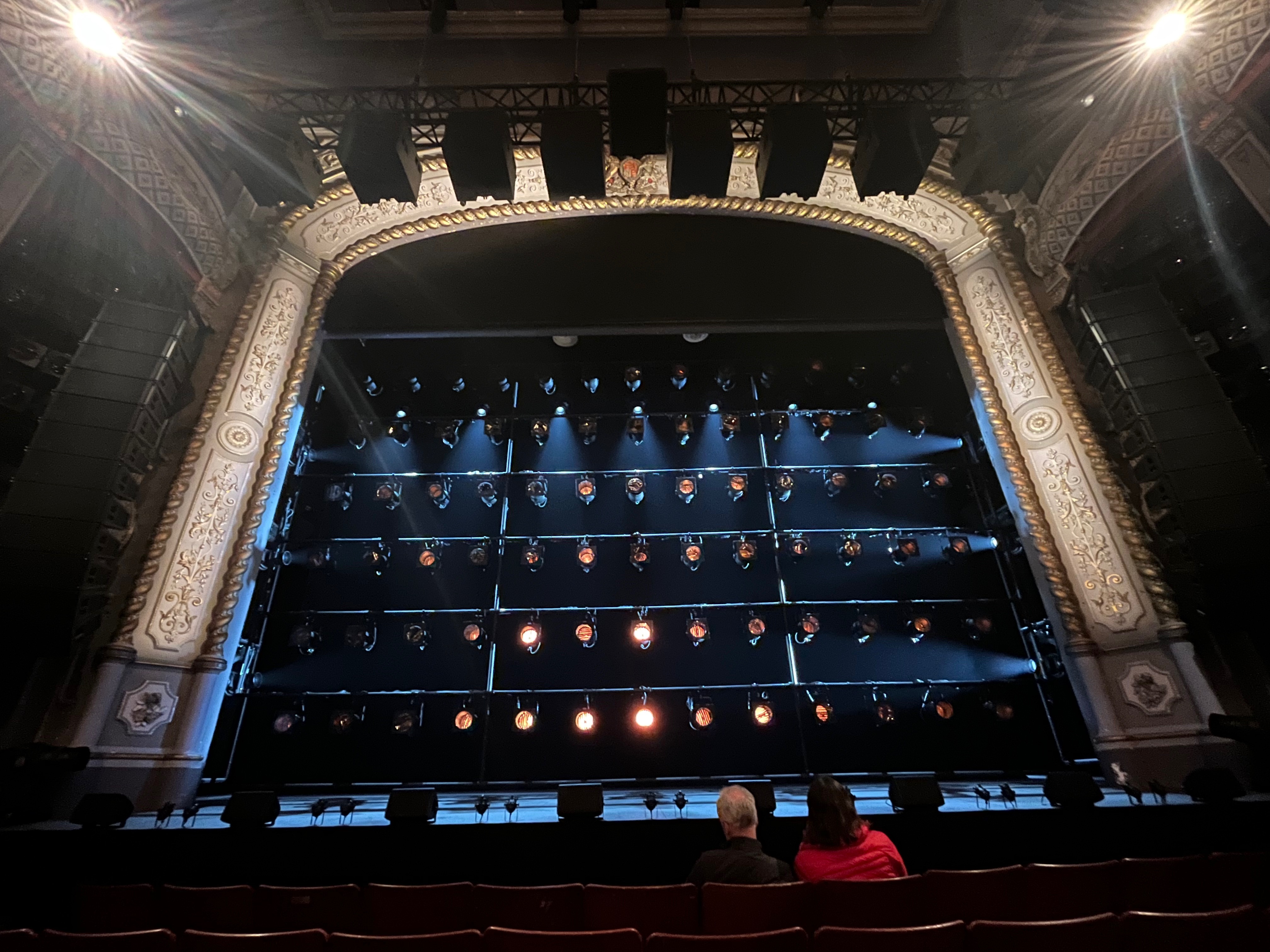 The stage before the start of Just For One Day at the Old Vic Theatre. A huge lighting rig, with 6 rows of lights, has been lowered in front of the stage, while the set is in darkness behind it.
