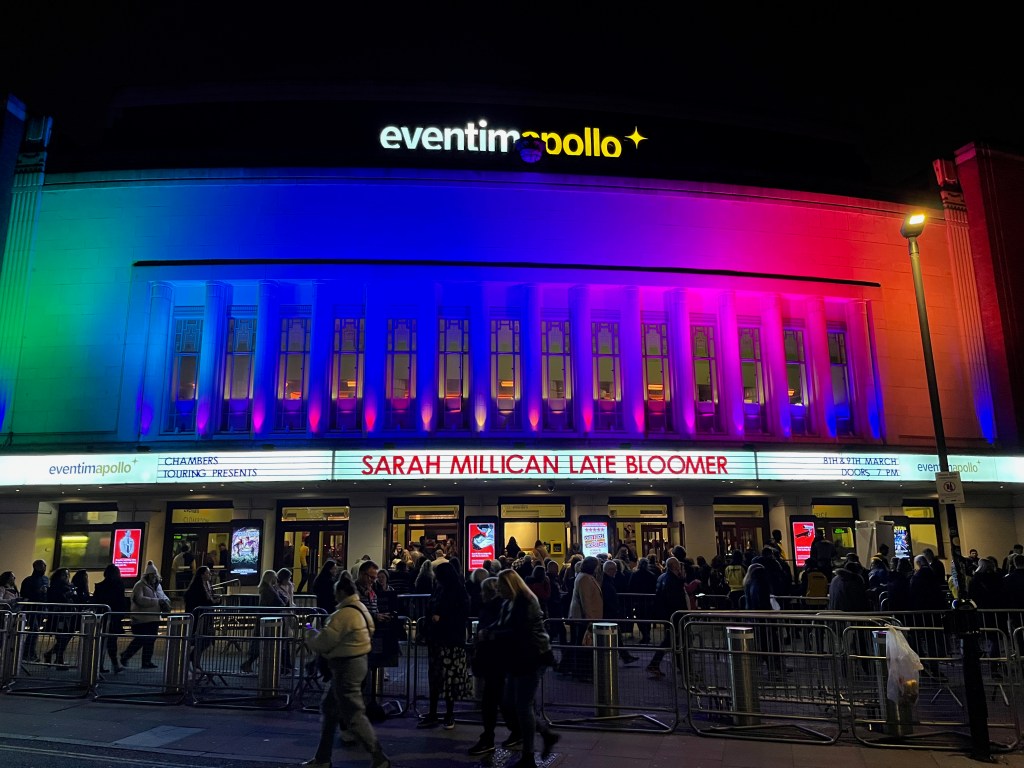The front of the Hammersmith Apollo theatre for Sarah Millican's show Late Bloomer. Animated rainbow colours illuminate the front of the building, so in this image the colours go from green on the left, through blue, pink and red, to orange on the right. Above the entrance a long white canopy has capital letters in the centre saying Sarah Millican Late Bloomer, along with small text on the left that says Chambers Touring Presents, and small text on the right that says 8th and 9th March, doors 7pm.