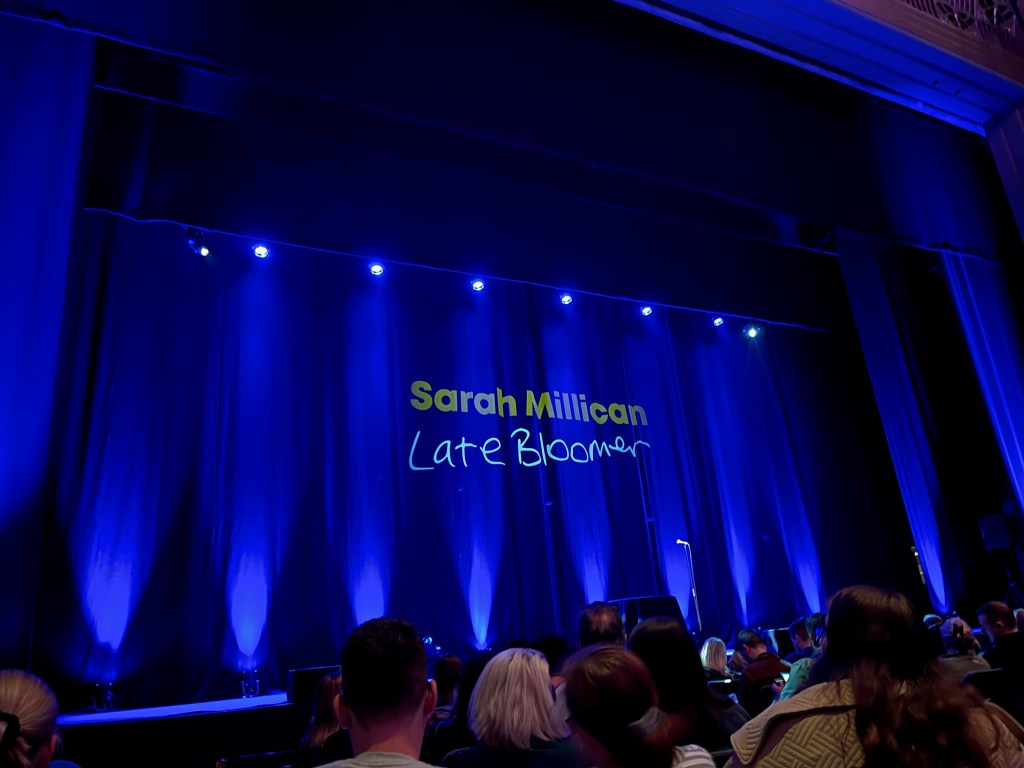 View towards the stage in the Hammersmith Apollo, which is lit up in blue with the title in large text on the back wall. The top line says Sarah Millican in green, typed letters, while the bottom line, Late Bloomer, is white and written like a child's handwriting, a bit like chalk on a blackboard.