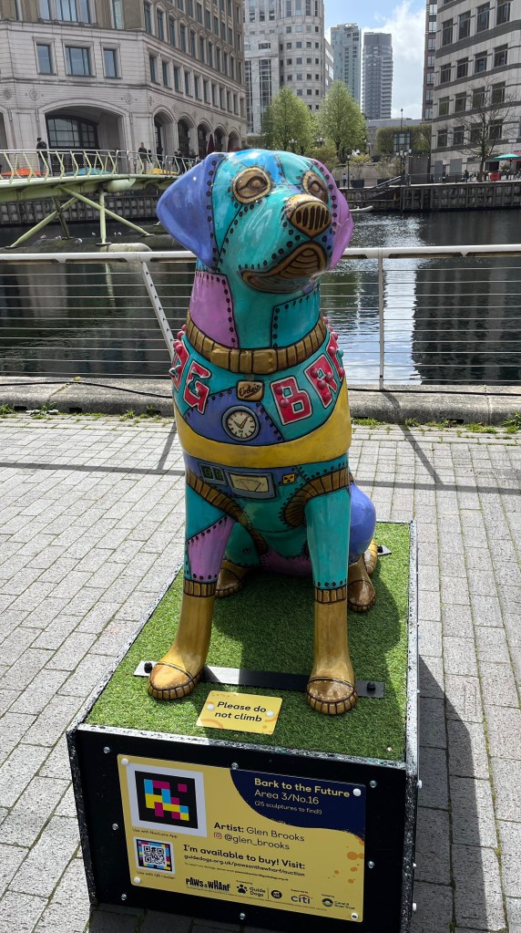 Front view of a sculpture of a multi-coloured metal futuristic patchwork robot guide dog. 'Dog Brown' is written across the dog's shoulders with rivets detailing the words in braille.