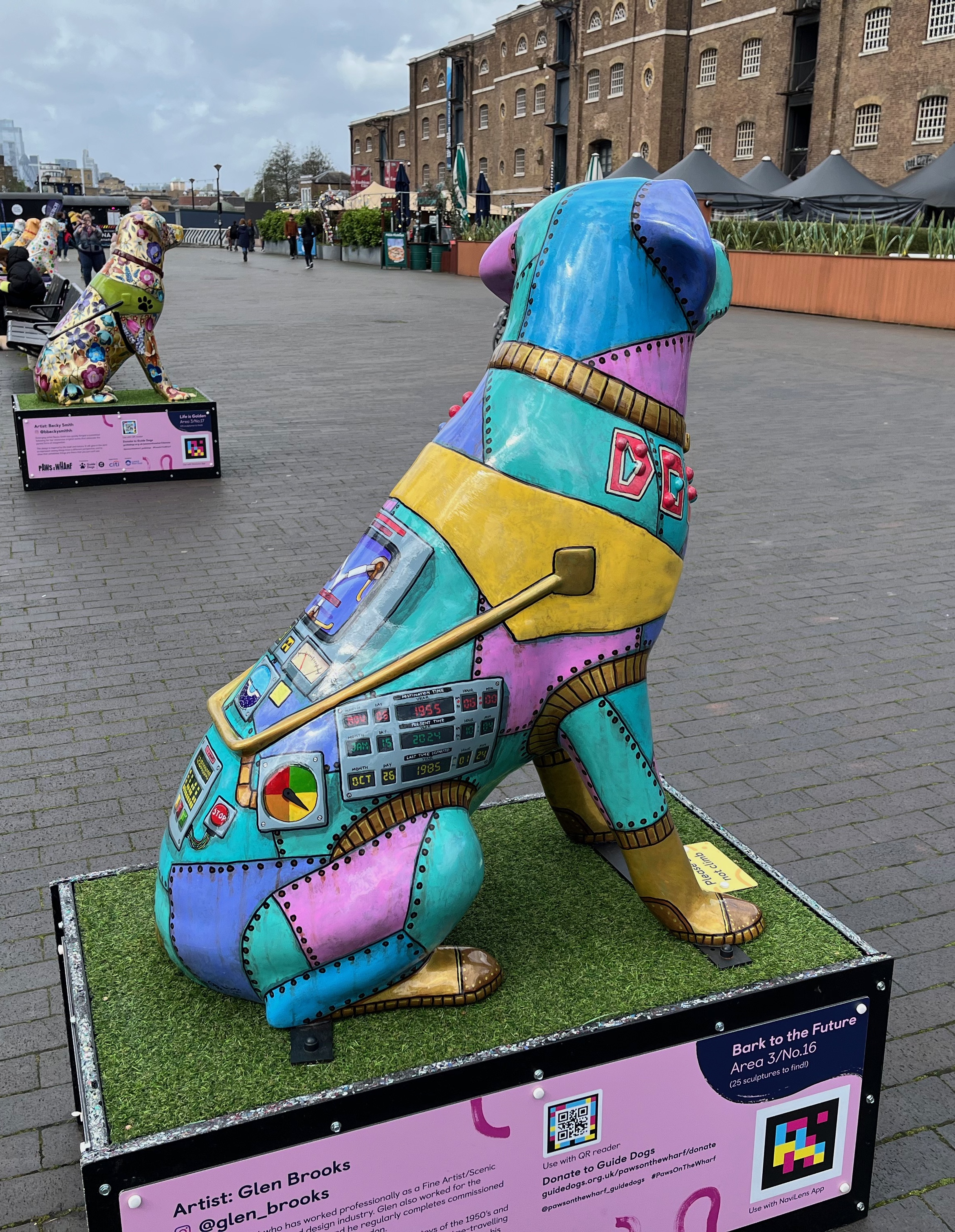 Right side view of a sculpture of a multi-coloured metal futuristic patchwork robot guide dog. Near the dog's right leg are 3 displays one above the other showing different date counters. The destination time is 6 June 1955 at 6am. The present time is 15 January 2024 at 10:40am. And the last time departed was 26 October 1985 at 1:24am.
